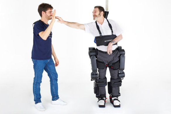 A man wearing an exoskeleton suit high-fives another man in casual attire, celebrating health and wellbeing against a plain white background.